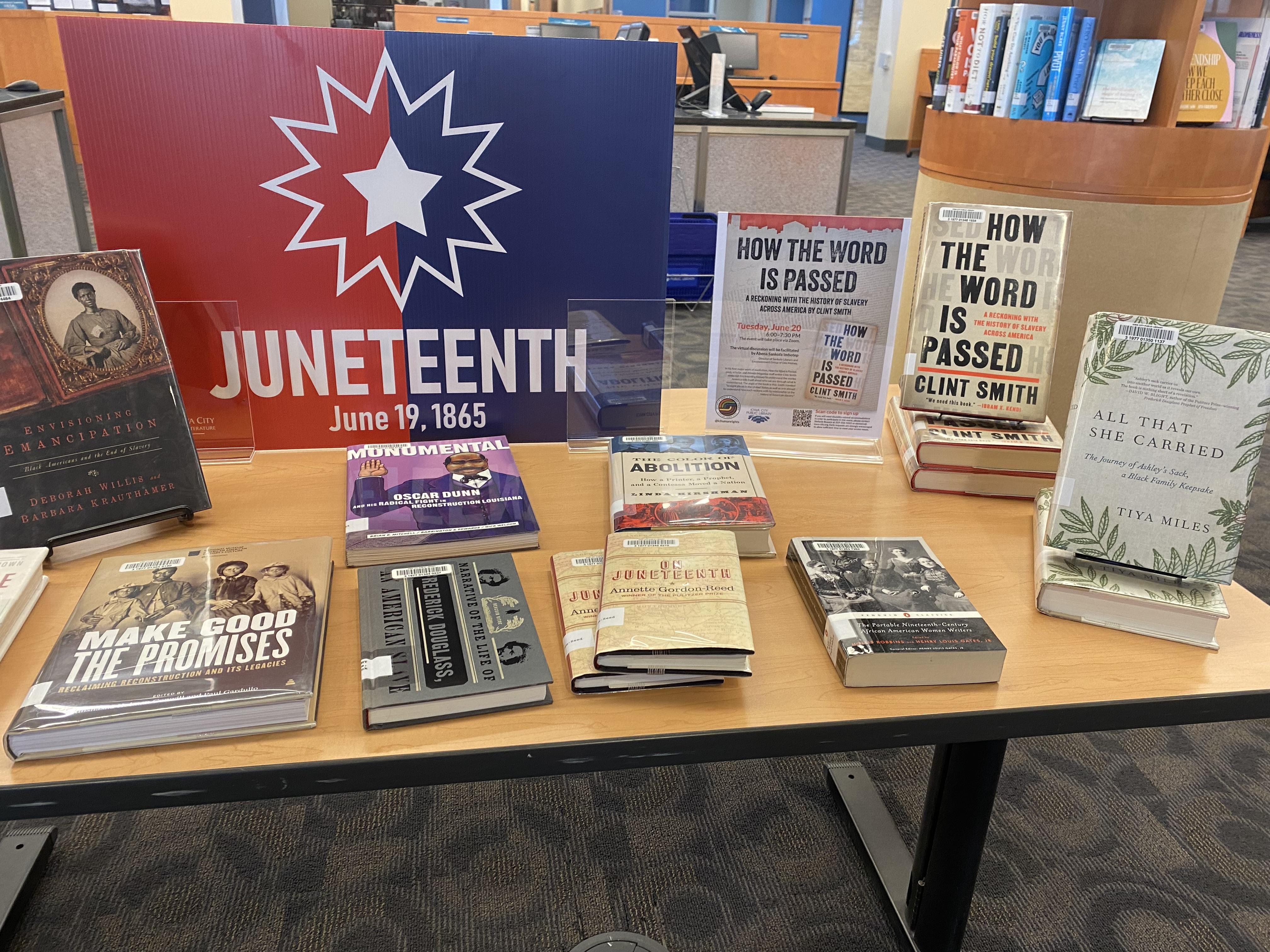 A book display with books about African-American history and contemporary topics, a red and white sign with a star that promotes the City of Iowa City's celebration of Juneteenth, and a promotion of a book discussion for "How the Word is Passed" by Clint Smith. 